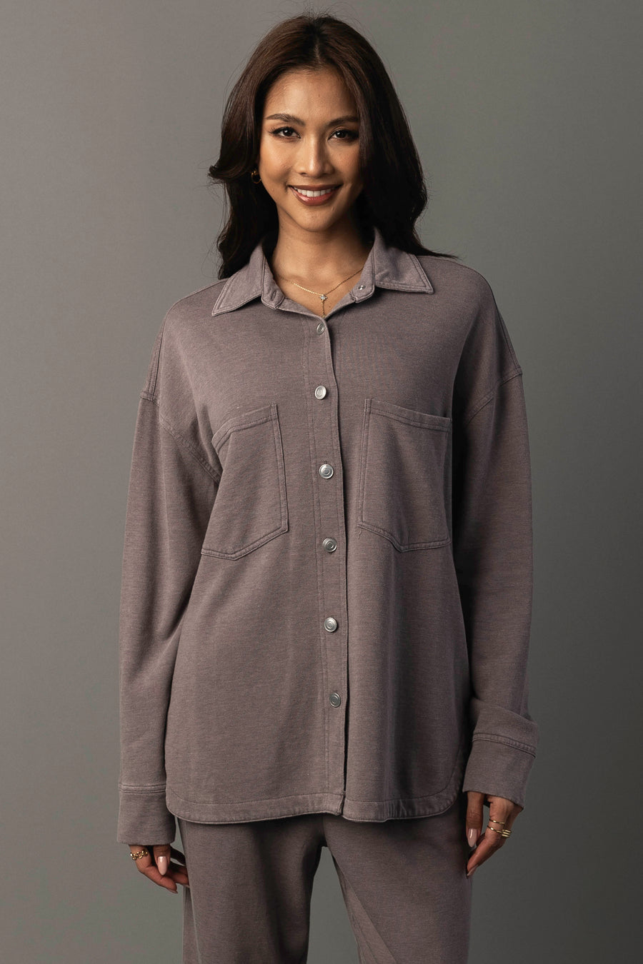 A woman with long, dark hair stands against a plain gray background, smiling as she wears the Panache Apparel Collection’s Gray Washed Soft Button Up and matching pants. The outfit features a loose fit in a muted mauve-gray tone, and her hands are relaxed at her sides.