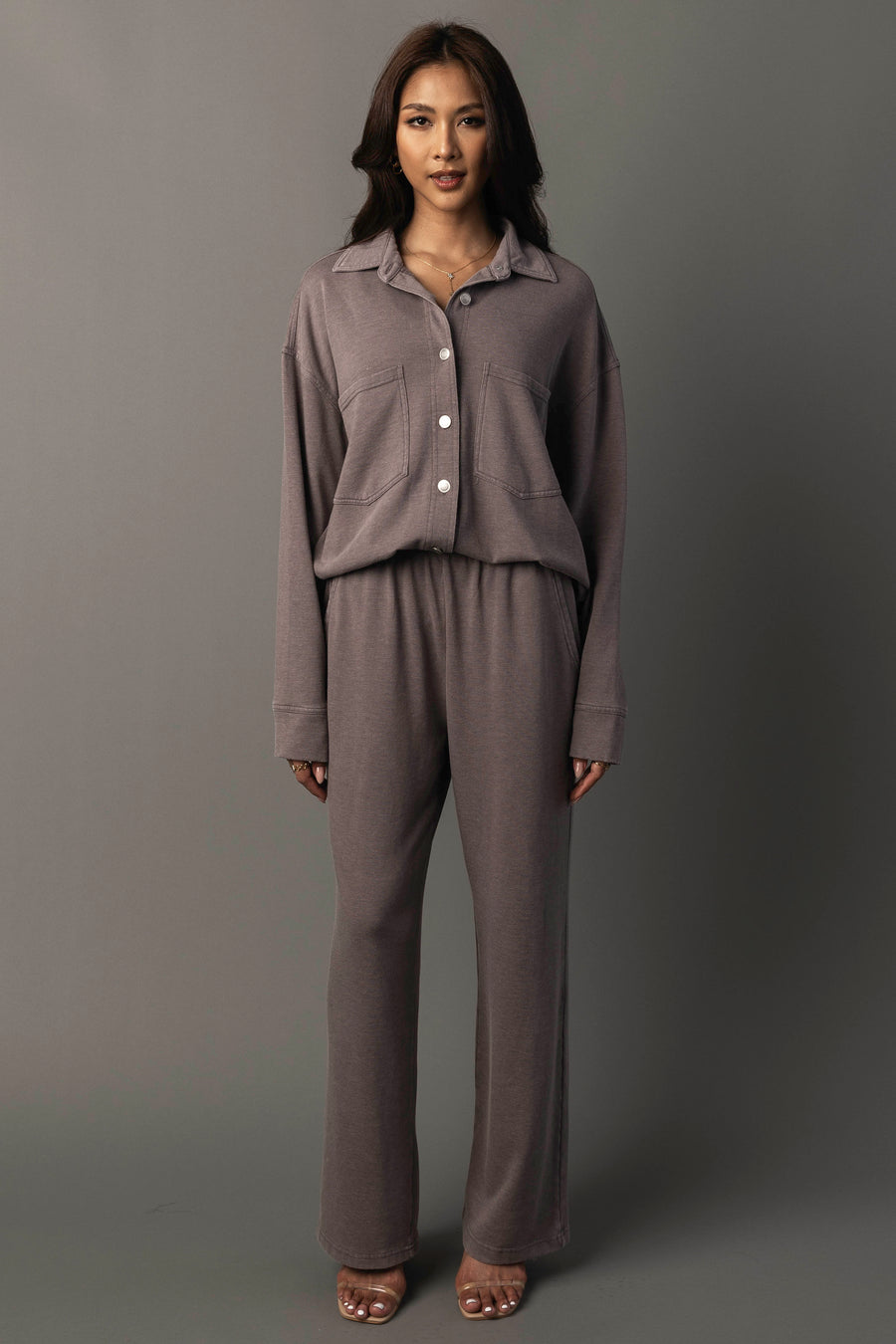 A woman stands against a gray background, wearing the Gray Washed Soft Button Up by Panache Apparel Collection. A loose-fitting, long-sleeve top, paired with matching high-waisted pants in a muted brown shade and open-toed heels.