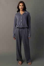 A woman stands smiling, dressed in the Panache Apparel Collection’s Slate Blue Washed Soft Button Up with matching wide-leg pants, her hands relaxed by her sides against a plain gray background.