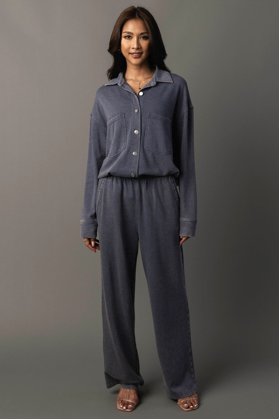 A woman stands smiling, dressed in the Panache Apparel Collection’s Slate Blue Washed Soft Button Up with matching wide-leg pants, her hands relaxed by her sides against a plain gray background.