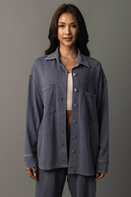 A woman with long dark hair wears the Panache Apparel Collection’s Slate Blue Washed Soft Button Up layered over a white top and blue pants, standing against a gray background and smiling gently at the camera.