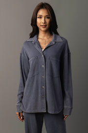 A woman with long dark hair is wearing the Panache Apparel Collection’s Slate Blue Washed Soft Button Up, a loose-fitting shirt with front pockets paired with matching pants, standing against a plain, neutral background.