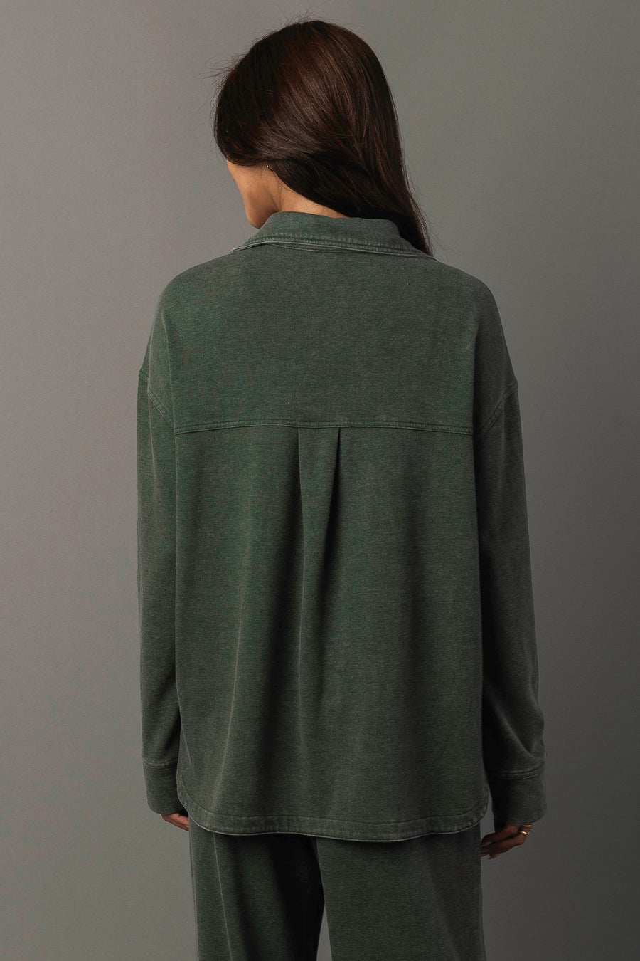 A person with long brown hair, wearing Panache Apparel Collection’s Pine Green Washed Soft Button Up and matching pants, stands facing away against a plain gray background.