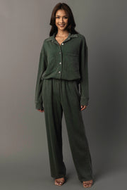A woman stands smiling, wearing the Panache Apparel Collection’s Pine Green Washed Soft Button Up with front pockets and matching wide-leg pants. She has long hair and pairs the outfit with open-toed heels against a plain gray background.