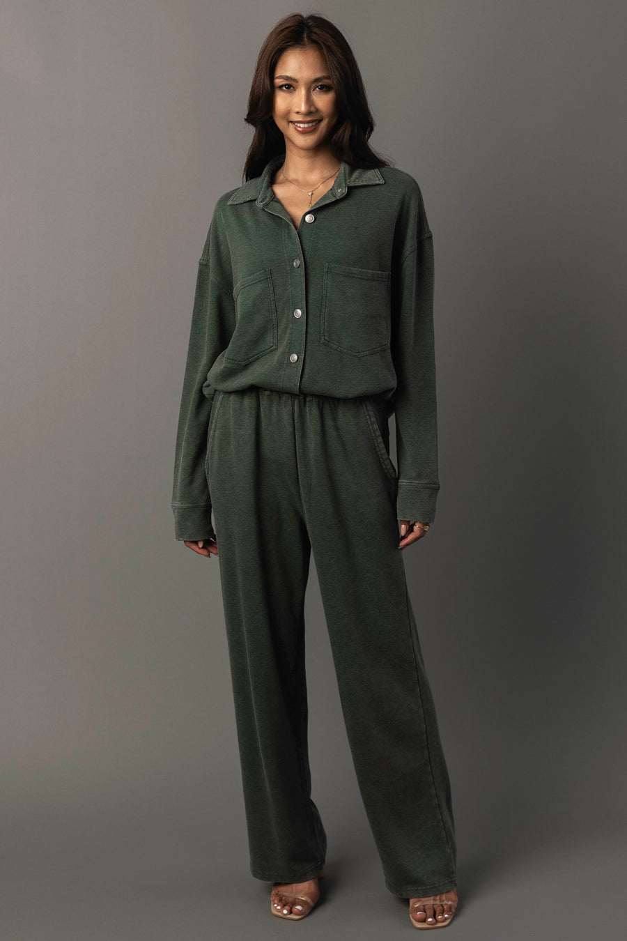 A woman stands smiling, wearing the Panache Apparel Collection’s Pine Green Washed Soft Button Up with front pockets and matching wide-leg pants. She has long hair and pairs the outfit with open-toed heels against a plain gray background.