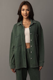 A woman with long dark hair stands against a plain gray background, smiling slightly as she wears the Panache Apparel Collection Pine Green Washed Soft Button Up over a white crop top and matching green pants.