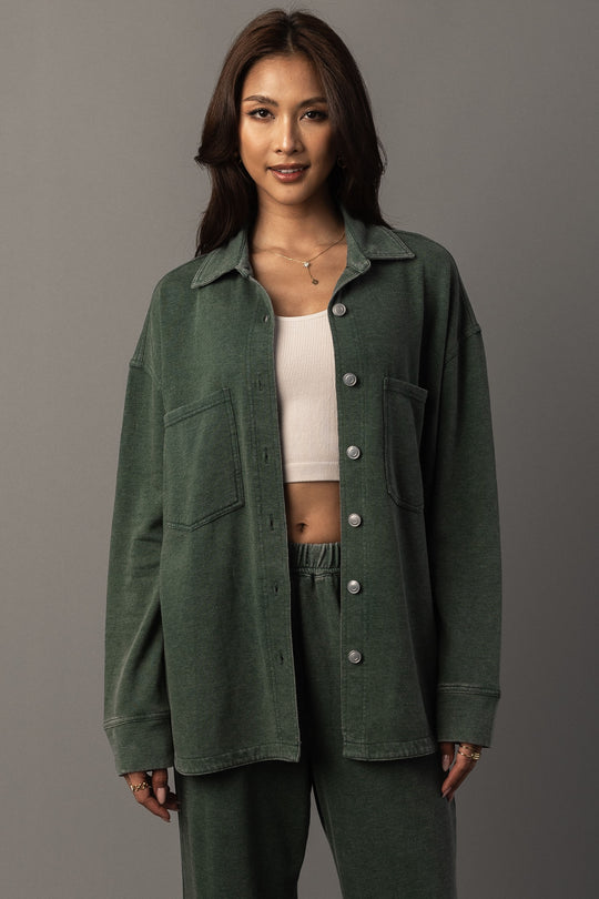 A woman with long dark hair stands against a plain gray background, smiling slightly as she wears the Panache Apparel Collection Pine Green Washed Soft Button Up over a white crop top and matching green pants.