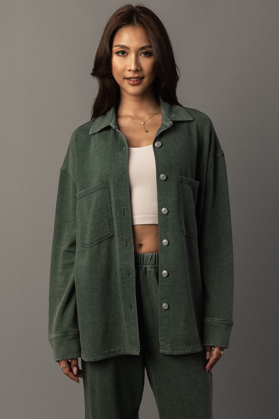 A woman with long dark hair stands against a plain gray background, smiling slightly as she wears the Panache Apparel Collection Pine Green Washed Soft Button Up over a white crop top and matching green pants.