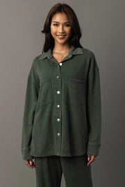 A woman with long brown hair is smiling and wearing the Panache Apparel Collection’s Pine Green Washed Soft Button Up with two large front pockets and matching pants, standing against a plain gray background.