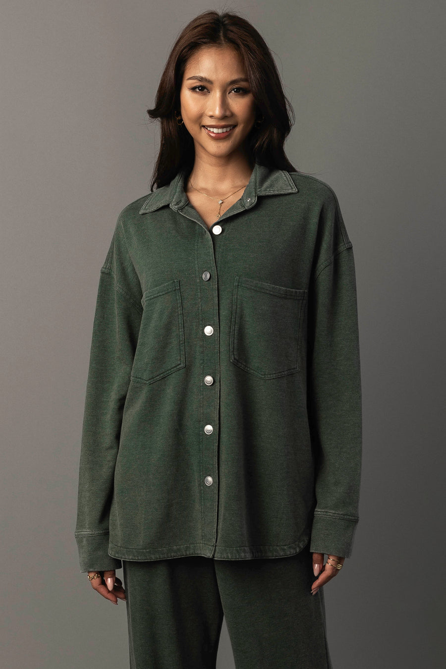 A woman with long brown hair is smiling and wearing the Panache Apparel Collection’s Pine Green Washed Soft Button Up with two large front pockets and matching pants, standing against a plain gray background.