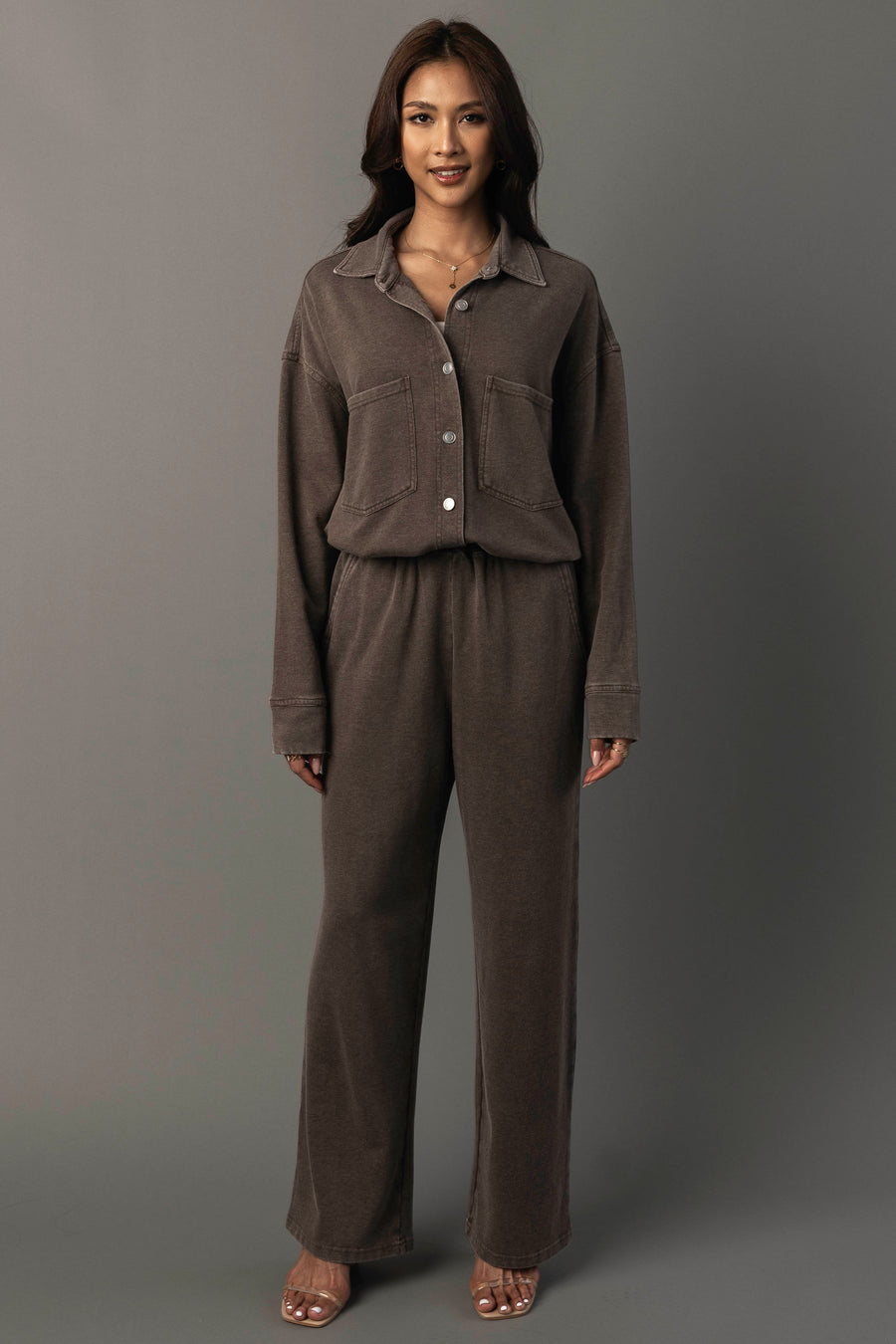A woman stands against a plain gray background, smiling with her arms relaxed by her sides, wearing Panache Apparel Collection’s Cocoa Washed Soft Button Up and matching wide-leg pants paired with open-toed heels.