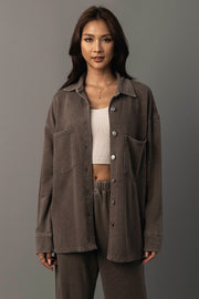 A woman with long dark hair wears the Cocoa Washed Soft Button Up from Panache Apparel Collection, styled over a white crop top and matching brown pants. She stands against a gray background, facing forward with a neutral expression.