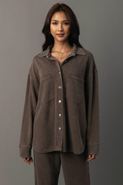 A woman with long dark hair models the Panache Apparel Collection’s Cocoa Washed Soft Button Up, a loose-fitting brown shirt with two chest pockets, paired with matching brown pants, standing against a plain gray background.