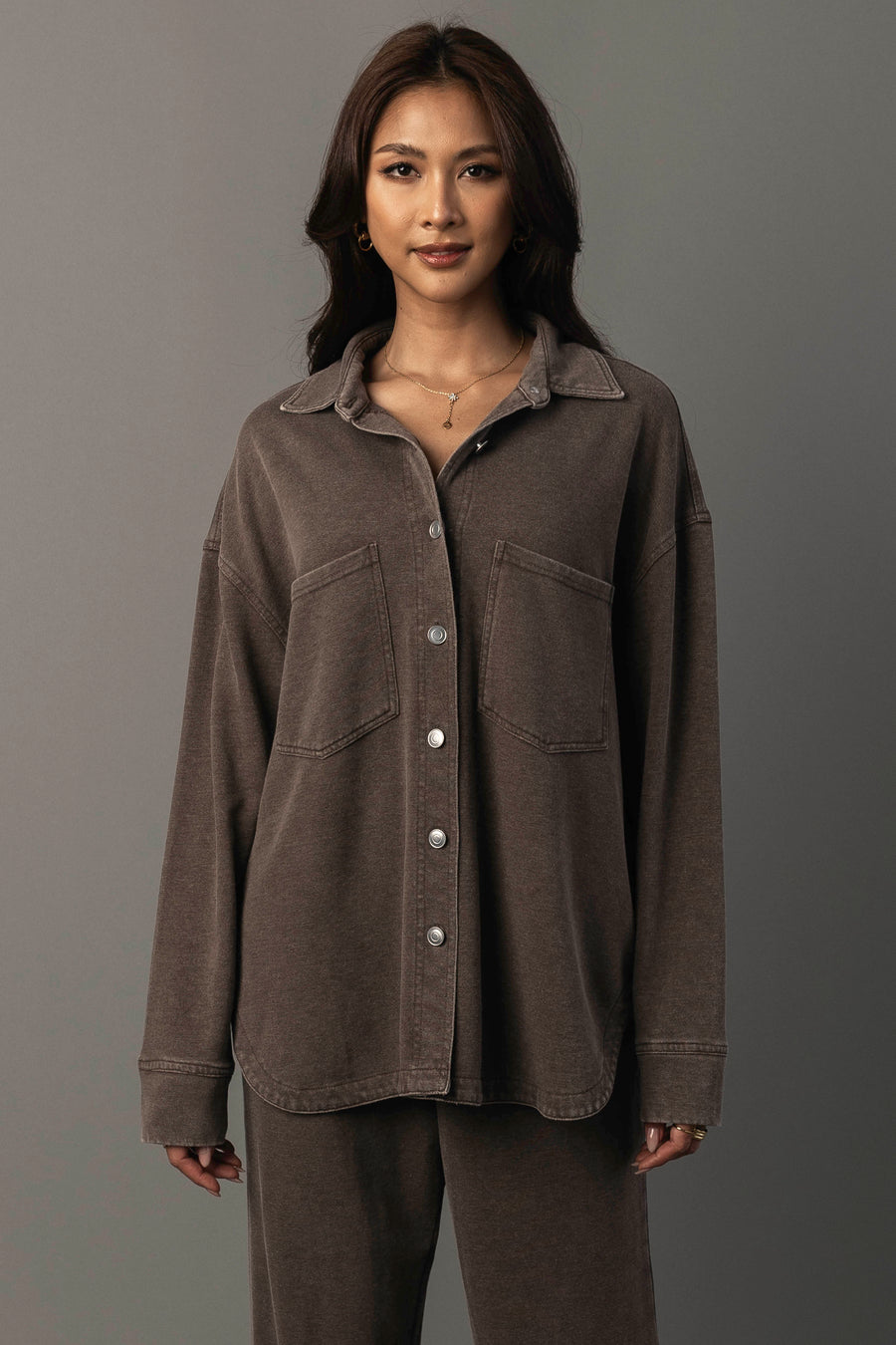 A woman with long dark hair models the Panache Apparel Collection’s Cocoa Washed Soft Button Up, a loose-fitting brown shirt with two chest pockets, paired with matching brown pants, standing against a plain gray background.