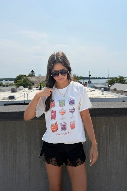 A woman with long hair and sunglasses stands on a rooftop with trees, buildings, and water in the background under a partly cloudy sky. She’s wearing the Panache Apparel Collection’s “Here For The Boos Tee”—a women’s Halloween t-shirt with colorful drinks—paired with black lace shorts.
