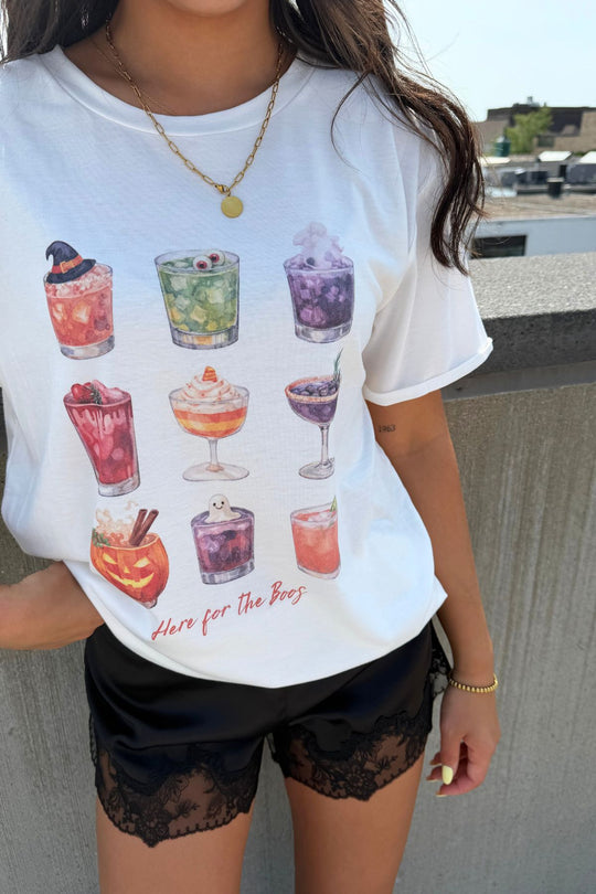 A woman stands outside by a concrete wall wearing the Panache Apparel Collection’s “Here For The Boos Tee,” which features colorful illustrated Halloween cocktails and the slogan “Here for the Boos.” She pairs it with black lace-trim shorts and gold jewelry.