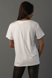 A person with long dark hair, wearing the Panache Apparel Collection “Coffee Is Always A Good Idea Tee” and black pants, stands facing away from the camera against a gray background.