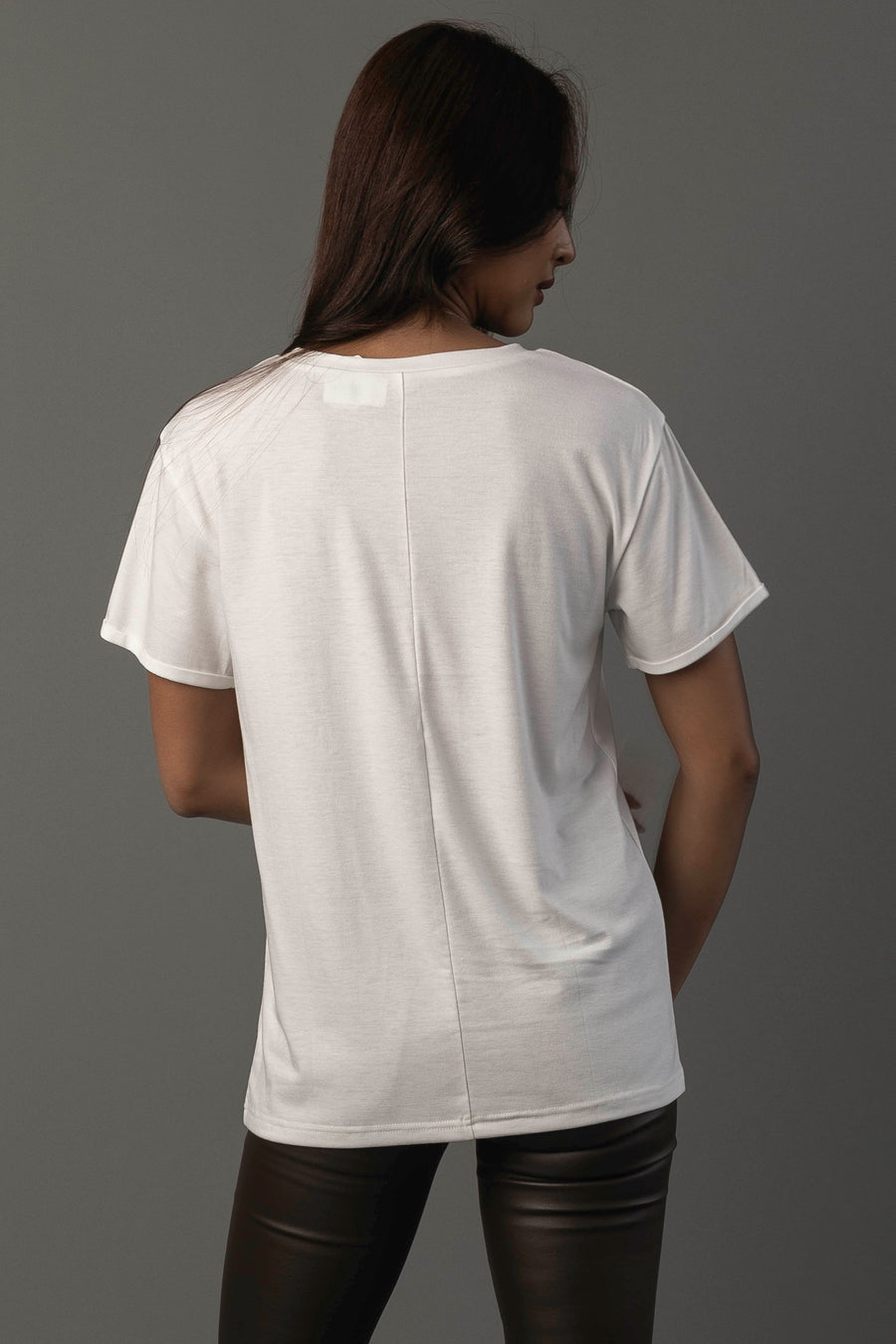 A person with long dark hair, wearing the Panache Apparel Collection “Coffee Is Always A Good Idea Tee” and black pants, stands facing away from the camera against a gray background.