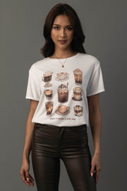A woman models the “Coffee Is Always A Good Idea Tee” by Panache Apparel Collection, featuring illustrations of various coffee drinks. She stands against a gray background, styled with dark brown pants and gold jewelry.