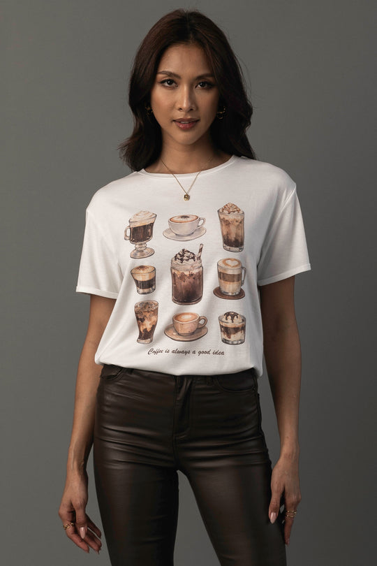 A woman models the “Coffee Is Always A Good Idea Tee” by Panache Apparel Collection, featuring illustrations of various coffee drinks. She stands against a gray background, styled with dark brown pants and gold jewelry.