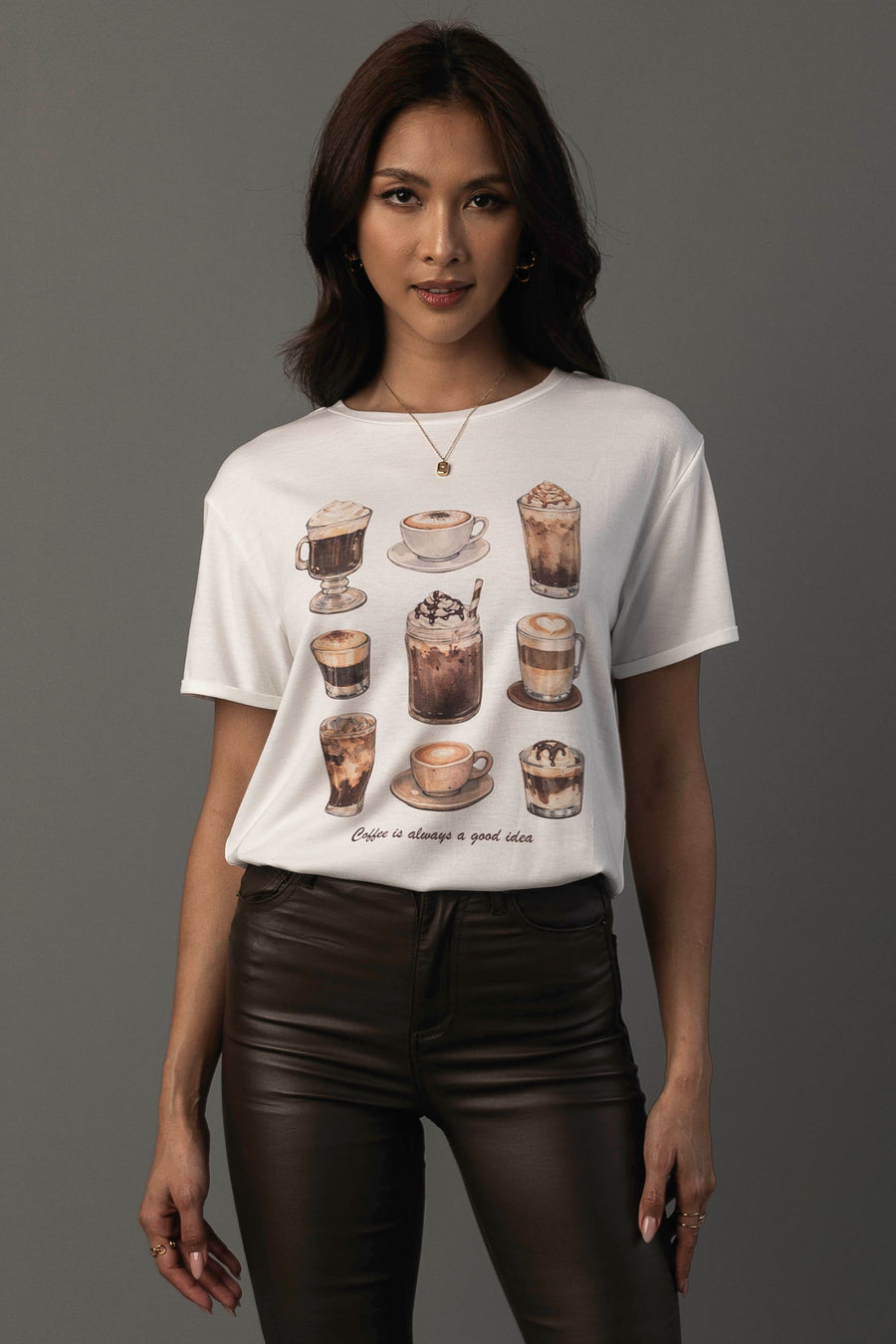 A woman models the “Coffee Is Always A Good Idea Tee” by Panache Apparel Collection, featuring illustrations of various coffee drinks. She stands against a gray background, styled with dark brown pants and gold jewelry.