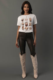 A woman with long, dark hair and a neutral expression stands against a gray background wearing the “Coffee Is Always A Good Idea Tee” by Panache Apparel Collection—a white T-shirt featuring drink illustrations—paired with black leather pants and tall beige cowboy boots.
