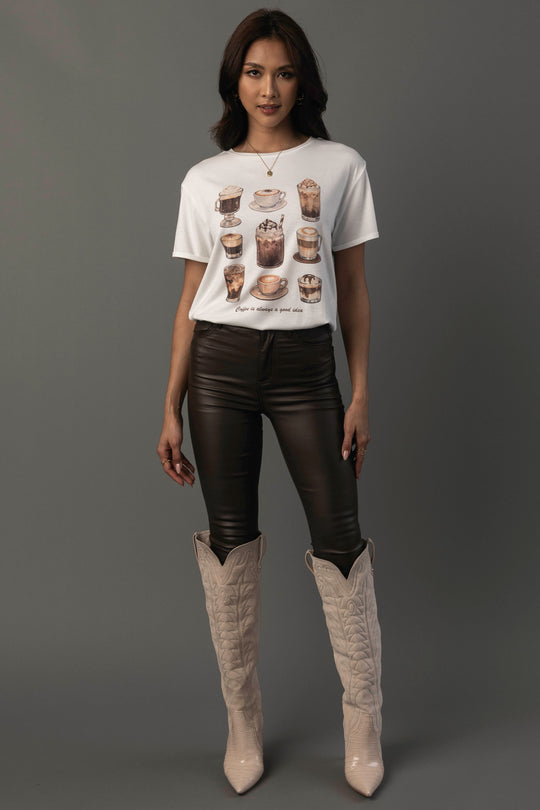 A woman with long, dark hair and a neutral expression stands against a gray background wearing the “Coffee Is Always A Good Idea Tee” by Panache Apparel Collection—a white T-shirt featuring drink illustrations—paired with black leather pants and tall beige cowboy boots.