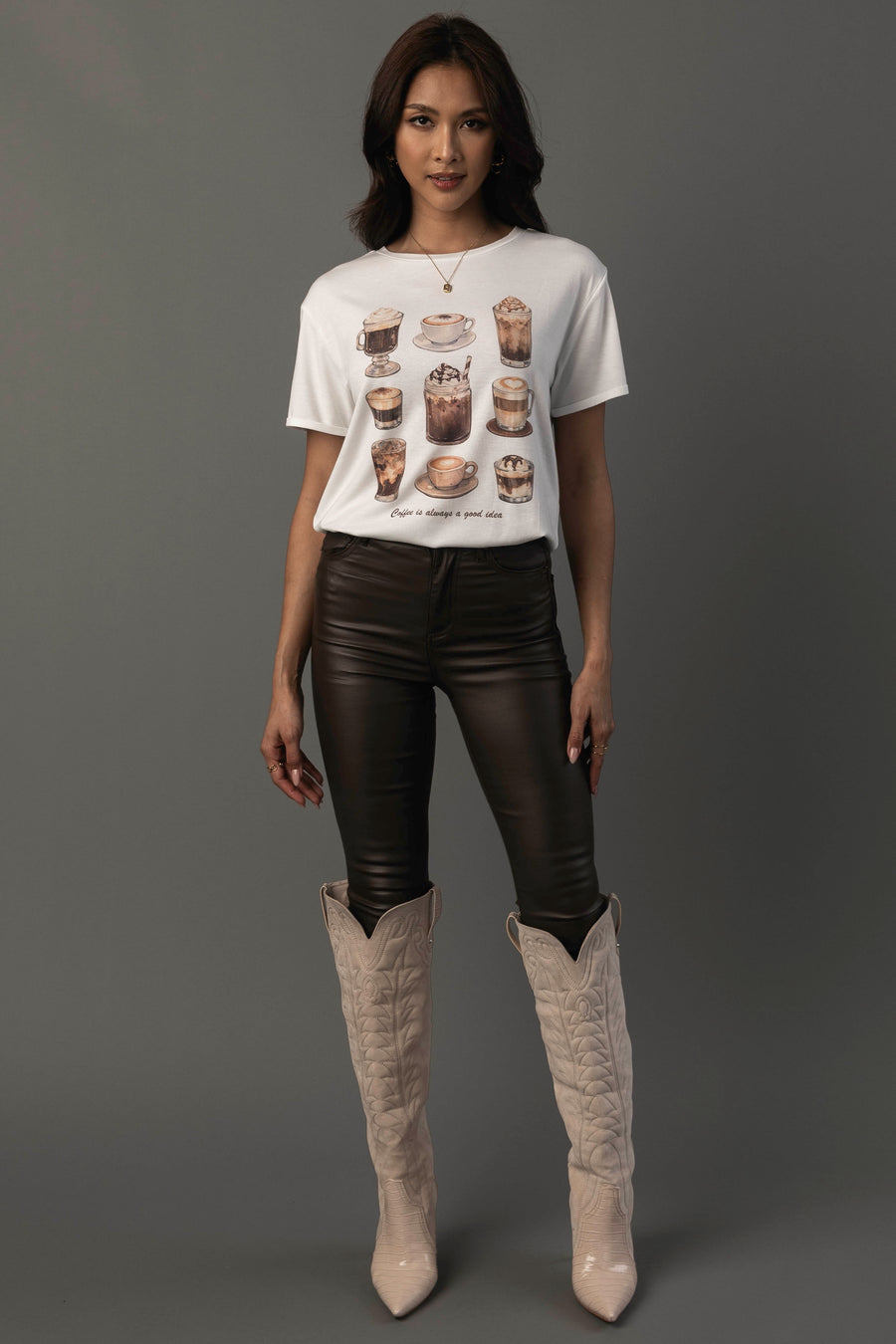 A woman with long, dark hair and a neutral expression stands against a gray background wearing the “Coffee Is Always A Good Idea Tee” by Panache Apparel Collection—a white T-shirt featuring drink illustrations—paired with black leather pants and tall beige cowboy boots.