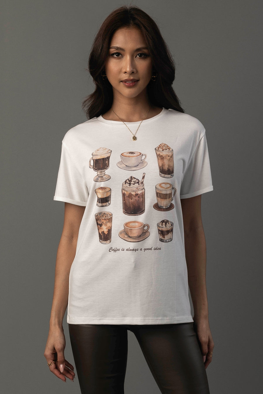 A woman stands against a plain gray background wearing the Panache Apparel Collection’s “Coffee Is Always A Good Idea Tee,” a white t-shirt featuring illustrations of coffee drinks and the phrase “Coffee is always a good idea.”.