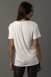 A woman with long brown hair wears the Panache Apparel Collection Whiskey Cocktails Tee, a plain white short-sleeve t-shirt, paired with black pants. She stands against a gray background, facing away from the camera.