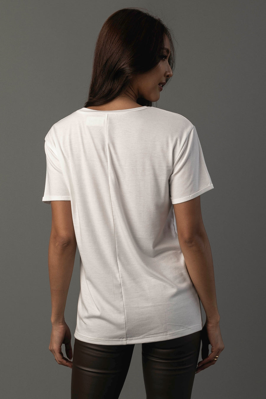 A woman with long brown hair wears the Panache Apparel Collection Whiskey Cocktails Tee, a plain white short-sleeve t-shirt, paired with black pants. She stands against a gray background, facing away from the camera.