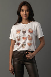 A woman with long dark hair wears the Whiskey Cocktails Tee by Panache Apparel Collection—a white t-shirt adorned with illustrations of various whiskey cocktails in glasses—paired with dark brown pants, standing against a plain gray background.