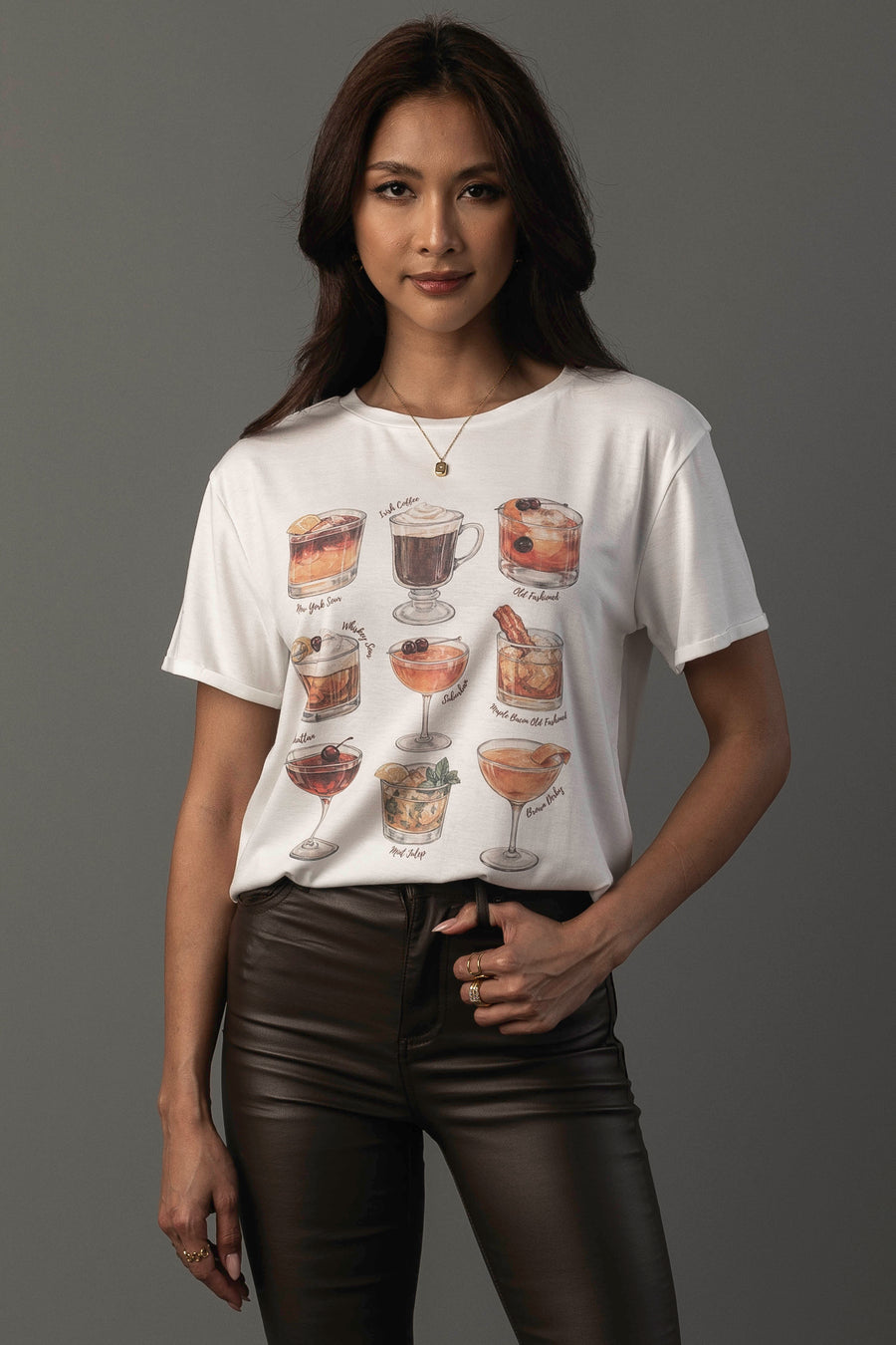 A woman with long dark hair wears the Whiskey Cocktails Tee by Panache Apparel Collection—a white t-shirt adorned with illustrations of various whiskey cocktails in glasses—paired with dark brown pants, standing against a plain gray background.