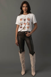 A woman with long dark hair and a slight smile stands against a gray background, wearing the Panache Apparel Collection’s Whiskey Cocktails Tee—a white t-shirt featuring drink illustrations—paired with black leather pants and tall beige cowboy boots. One hand rests on her hip.
