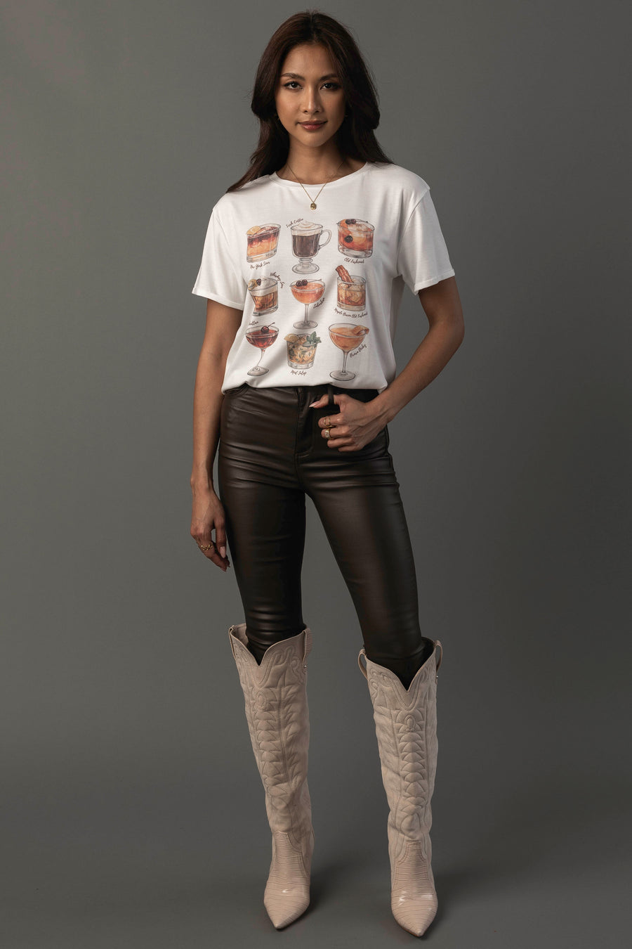 A woman with long dark hair and a slight smile stands against a gray background, wearing the Panache Apparel Collection’s Whiskey Cocktails Tee—a white t-shirt featuring drink illustrations—paired with black leather pants and tall beige cowboy boots. One hand rests on her hip.
