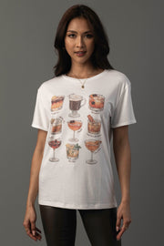 A woman wearing the Panache Apparel Collection’s Whiskey Cocktails Tee, featuring illustrations and names of various cocktails, stands against a gray background.