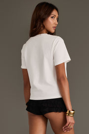 A woman stands facing away, looking over her shoulder. She wears the Unbothered Graphic Tee by Panache Apparel Collection, paired with black ruffled shorts and gold bracelets, against a plain brown background.