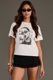 Woman wearing sunglasses, gold jewelry, and black ruffled shorts stands against a plain background, styled in the Unbothered Graphic Tee from Panache Apparel Collection featuring the phrase “I don’t care” over a black-and-white face.