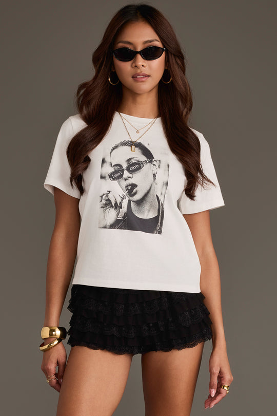 Woman wearing sunglasses, gold jewelry, and black ruffled shorts stands against a plain background, styled in the Unbothered Graphic Tee from Panache Apparel Collection featuring the phrase “I don’t care” over a black-and-white face.