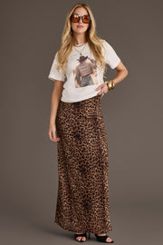 A woman wears sunglasses, gold jewelry, and black sandals, styled with a long leopard print skirt and the Panache Apparel Collection’s Howdy Western Graphic Tee—an off-white, 100% cotton shirt featuring a subtle cowboy graphic. She stands effortlessly comfortable against a plain brown background.