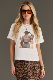 A woman stands against a plain gray background, wearing sunglasses, gold jewelry, and the Howdy Western Graphic Tee from Panache Apparel Collection, featuring a graphic of a person in a fringed jacket with “Howdy Honey” text, paired with a leopard print skirt.