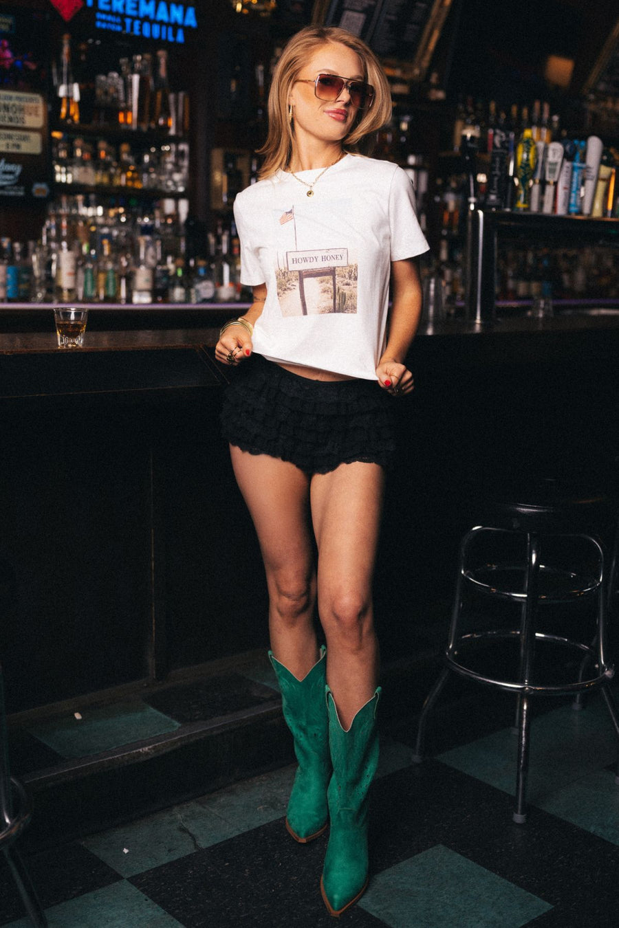 A woman in sunglasses, the Panache Apparel Collection Western Desert Graphic Tee in off white, black ruffled shorts, and green cowboy boots stands in a bar with bottles on shelves behind her and a drink on the counter.
