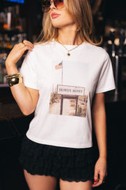 A woman wearing the Panache Apparel Collection’s Western Desert Graphic Tee—an off-white tee featuring a printed photo of a “Howdy Honey” sign and an American flag—paired with black ruffled shorts. She stands in a dimly lit bar.