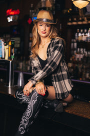 A woman wearing a cowboy hat, black top, and embroidered boots sits on a bar counter with a relaxed expression, surrounded by bottles and bar taps. She is dressed in the Panache Apparel Collection Tan & Black Plaid Button Up.