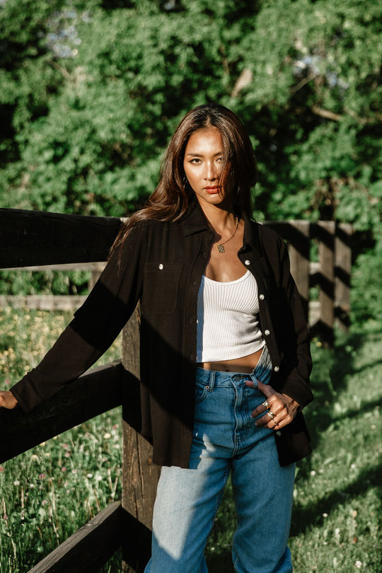 Outdoor lifestyle view of a woman wearing Panache Apparel’s black Super Soft button up open over a white ribbed top with blue jeans.