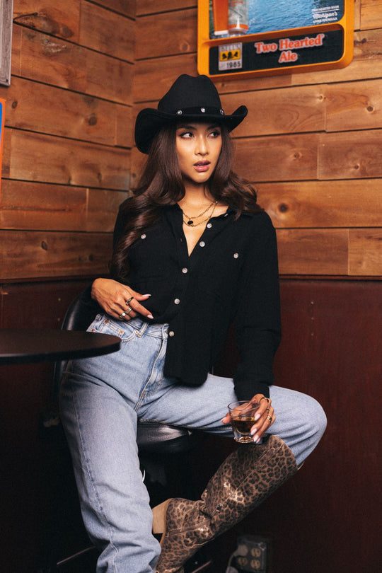 A woman wearing the Black Super Soft Button Up from Panache Apparel Collection, paired with jeans, a black cowboy hat, and leopard print boots, sits on a chair in a wood-paneled bar holding a drink and gazing to the side.