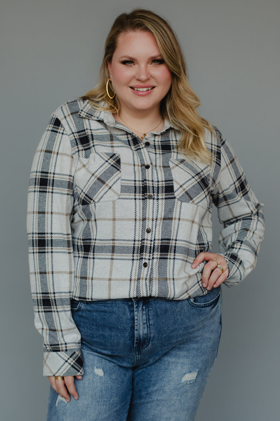 A woman with blonde hair wearing gold hoop earrings, a super soft Panache Apparel Collection Black, Light Gray & Brown Plaid Button Up and blue jeans smiles at the camera against a plain gray background.