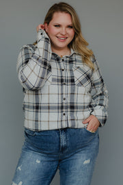 A smiling person with long blonde hair wears the Panache Apparel Collection’s Black, Light Gray & Brown Plaid Button Up and blue distressed jeans, standing against a plain gray background with one hand touching their hair.