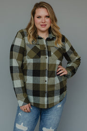 A person with long blonde hair stands with one hand on their hip against a plain gray background, wearing the Panache Apparel Collection Olive & Black Plaid Button Up a super soft, long-sleeved shirt paired with distressed blue jeans for a comfortable and casual look.