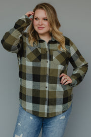 A woman stands against a gray background, touching her hair and smiling slightly, while wearing the Panache Apparel Collection’s Olive & Black Plaid Button Up paired with ripped blue jeans.